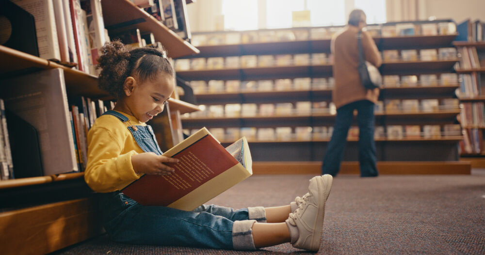 Little girl sitting on the floor in a library reading a big book
